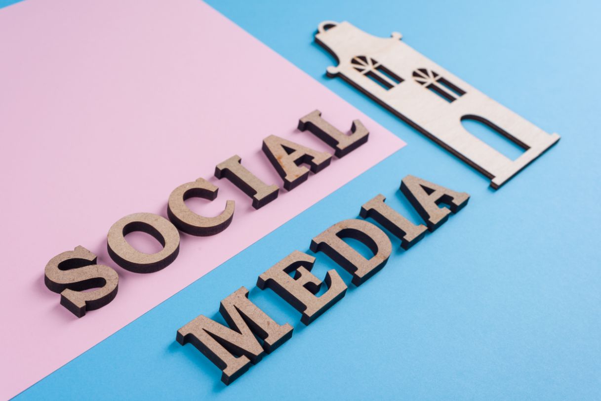 Wooden blocks spelling "social media" placed next to a wooden church on a blue and pink background.