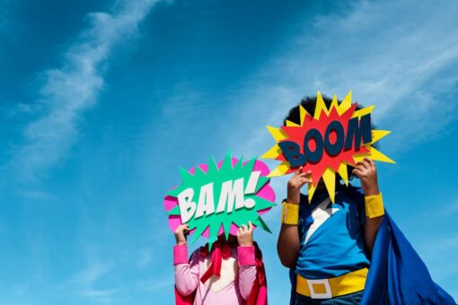 Children in superhero costumes holding signs in front of their faces saying bam! and boom! with the sky as the background